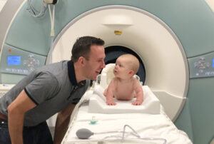 Father smiles at baby who is laying on the stretcher of an MRI machine